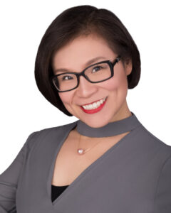 Headshot of a woman with short dark hair, wearing glasses and red lipstick. She chose a solid grey dress as the main component of her photoshoot wardrobe.